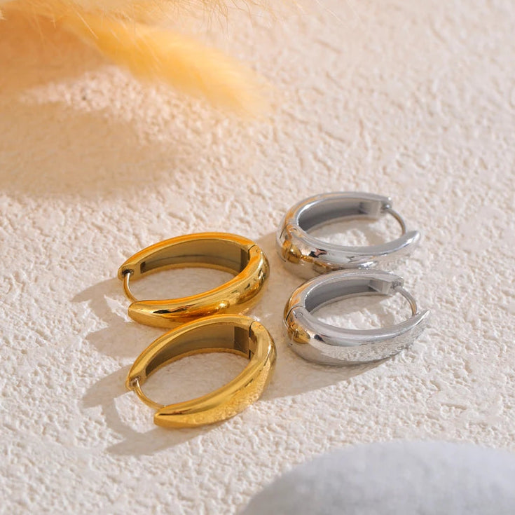 Two pairs of chunky hoop earrings laid flat on cream textured surface, gold pair on left and silver pair on right, mirror-polished tube hoops with hinged clasps, orange feather and white stone in background, warm angled light
