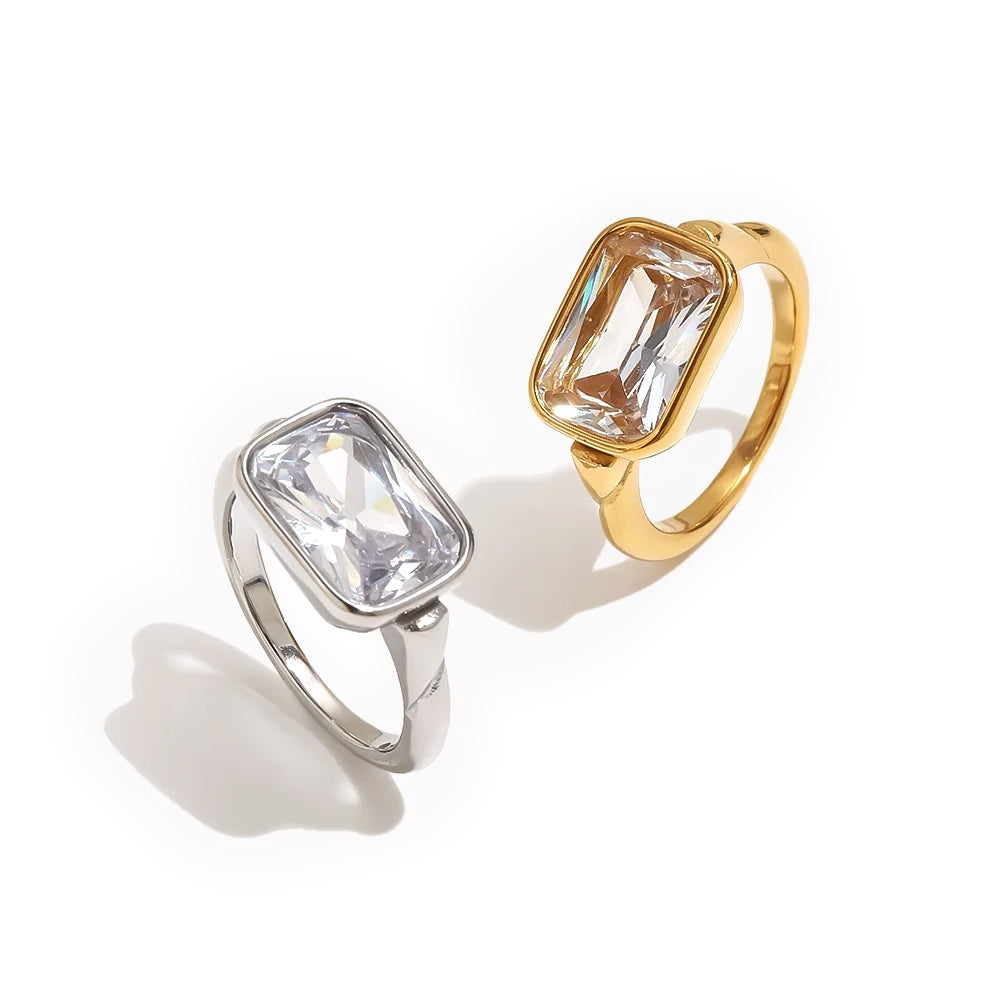 Gold and silver emerald cut stone bezel rings on white background, large clear faceted rectangular stone set flush in rounded bezel, both colorways shown together with soft warm shadow beneath
