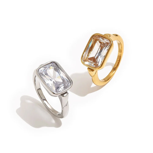 Gold and silver emerald cut stone bezel rings on white background, large clear faceted rectangular stone set flush in rounded bezel, both colorways shown together with soft warm shadow beneath
