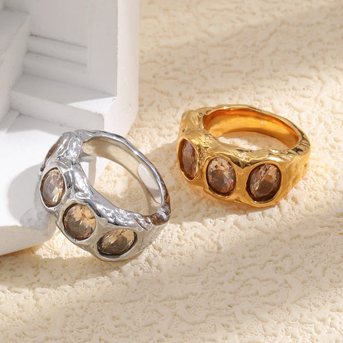Gold and silver hammered oval stone cocktail rings on cream textured surface, four champagne bezel-set stones facing up, white geometric object in background
