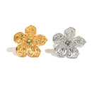 Gold and silver hammered texture flower rings side by side, five-petal sculptural bloom with green stone center, white background
