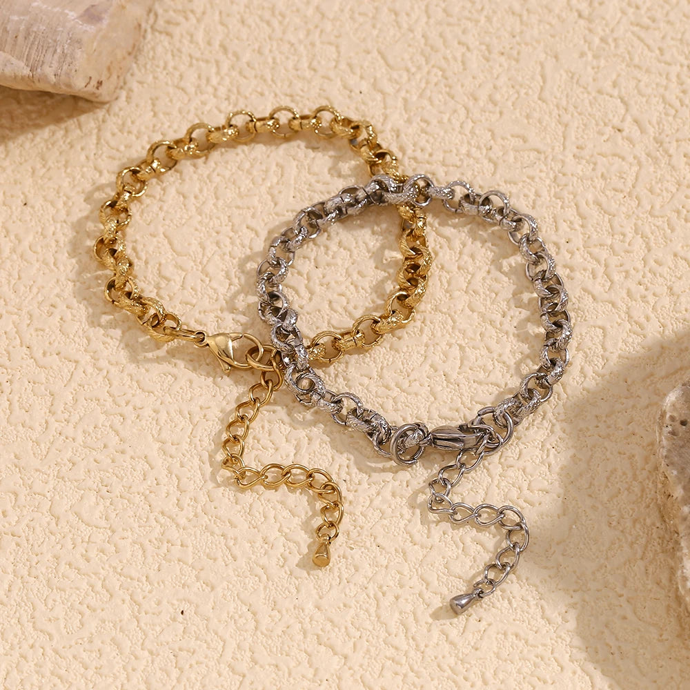 Gold and silver textured rolo chain bracelets overlapping on cream textured stone surface, hammered round links of both colorways shown together with lobster clasps and extender chains, warm angled light
