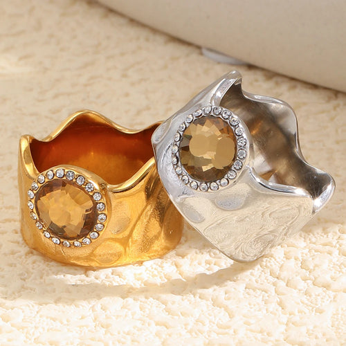 Gold and silver wavy wide band rings on cream knit surface, interior band visible, amber crystal halo detail
