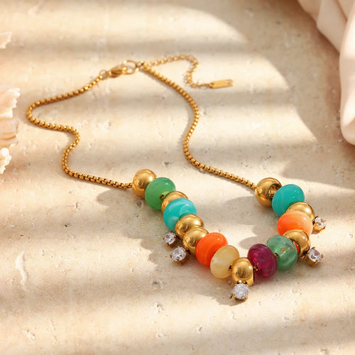 Gold colorful resin bead station necklace laid flat on warm sandy stone surface, multicolor resin beads and gold spheres with five clear stone stations visible, lobster clasp and extender chain, white shell in upper left corner
