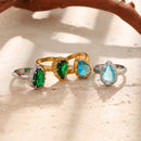 Four teardrop stone halo cocktail rings scattered on sandy stone surface with driftwood, gold band green and blue teardrop stones in center, silver band green and blue versions on sides, bright directional sunlight casting shadows
