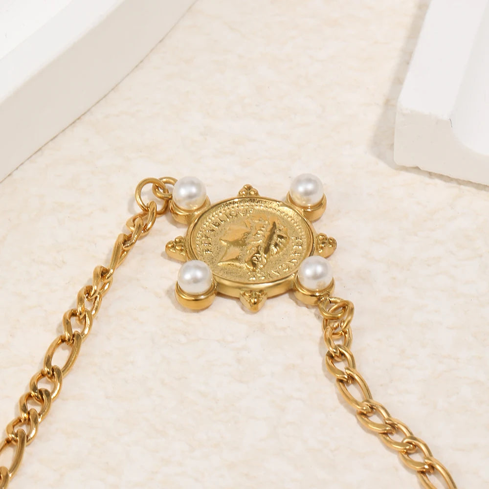 Close-up of gold pearl coin charm bracelet on cream fabric surface, reverse side of engraved coin showing classical relief figure with four pearl accents at corners, Figaro chain links on both sides, white jewelry box in background
