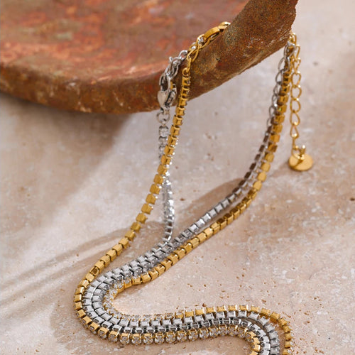 Gold and silver round stone tennis necklaces hanging over edge of terracotta stone block on cream surface, full length of both necklaces shown with continuous prong-set stones, extender chains and round tags visible, warm directional light
