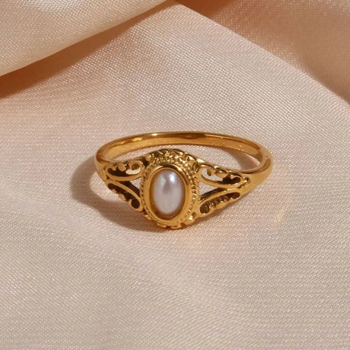 Gold filigree freshwater pearl ring resting on champagne satin fabric, oval pearl with soft warm luster inside beaded bezel and open scrollwork setting, fine band, warm ambient light
