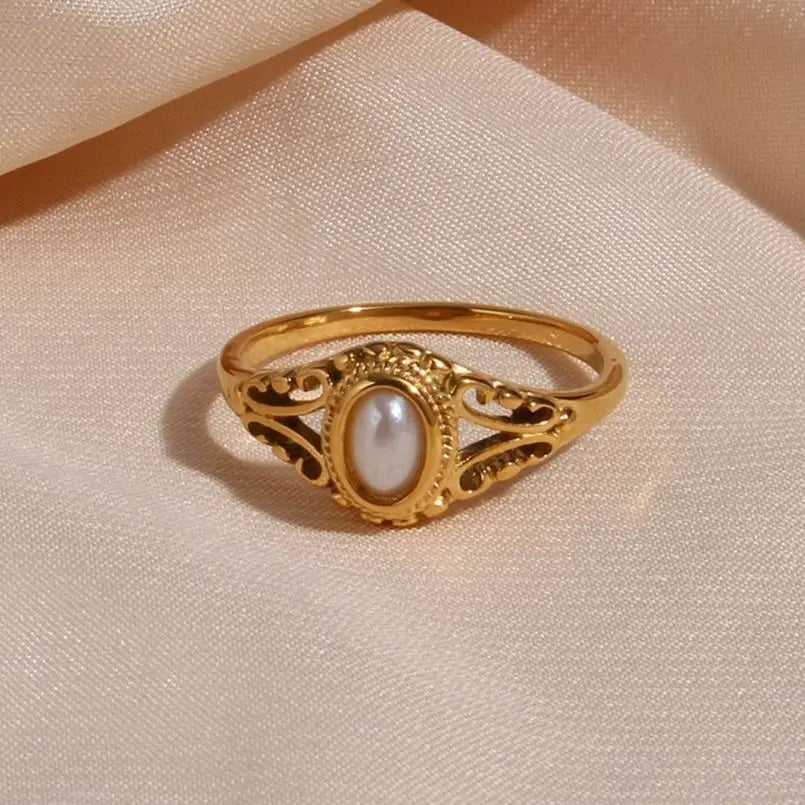 Gold filigree freshwater pearl ring resting on champagne satin fabric, oval pearl with soft warm luster inside beaded bezel and open scrollwork setting, fine band, warm ambient light
