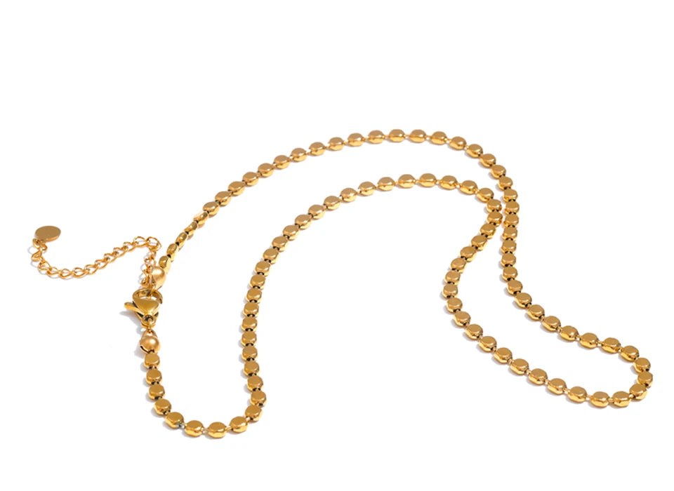 Gold disc chain necklace on white background, full length view showing lobster clasp with extender chain and round tag pendant at lower left, continuous flat disc links with mirror-polished faces, warm light with long soft shadow
