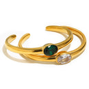 Gold oval stone open cuff bracelets in emerald green and clear stacked on white background, smooth high-polish band and bezel-set oval stones visible, open back cuff silhouette, warm shadow

