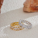 Gold and silver heart-cut eternity rings lying flat on textured cream fabric beside rose quartz stone, heart-shaped stones in four-prong settings visible across both bands, warm natural light

