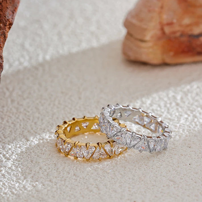 Gold and silver heart-cut eternity rings lying flat on textured cream fabric beside rose quartz stone, heart-shaped stones in four-prong settings visible across both bands, warm natural light

