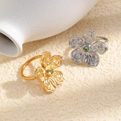Gold hammered texture flower ring on cream textured surface, angled view showing sculptural petals and green stone center, warm soft light
