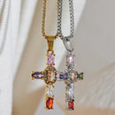 Gold and silver multicolor stone cross pendant necklaces hanging side by side on cream fabric background, both pendants showing colored oval stones and pavé center detail, gold and silver box chains, palm leaf shadow in background
