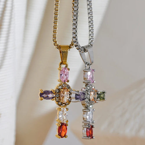 Gold and silver multicolor stone cross pendant necklaces hanging side by side on cream fabric background, both pendants showing colored oval stones and pavé center detail, gold and silver box chains, palm leaf shadow in background
