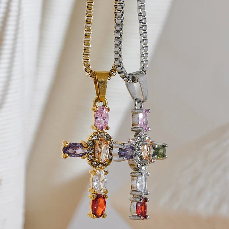 Gold and silver multicolor stone cross pendant necklaces hanging side by side on cream fabric background, both pendants showing colored oval stones and pavé center detail, gold and silver box chains, palm leaf shadow in background
