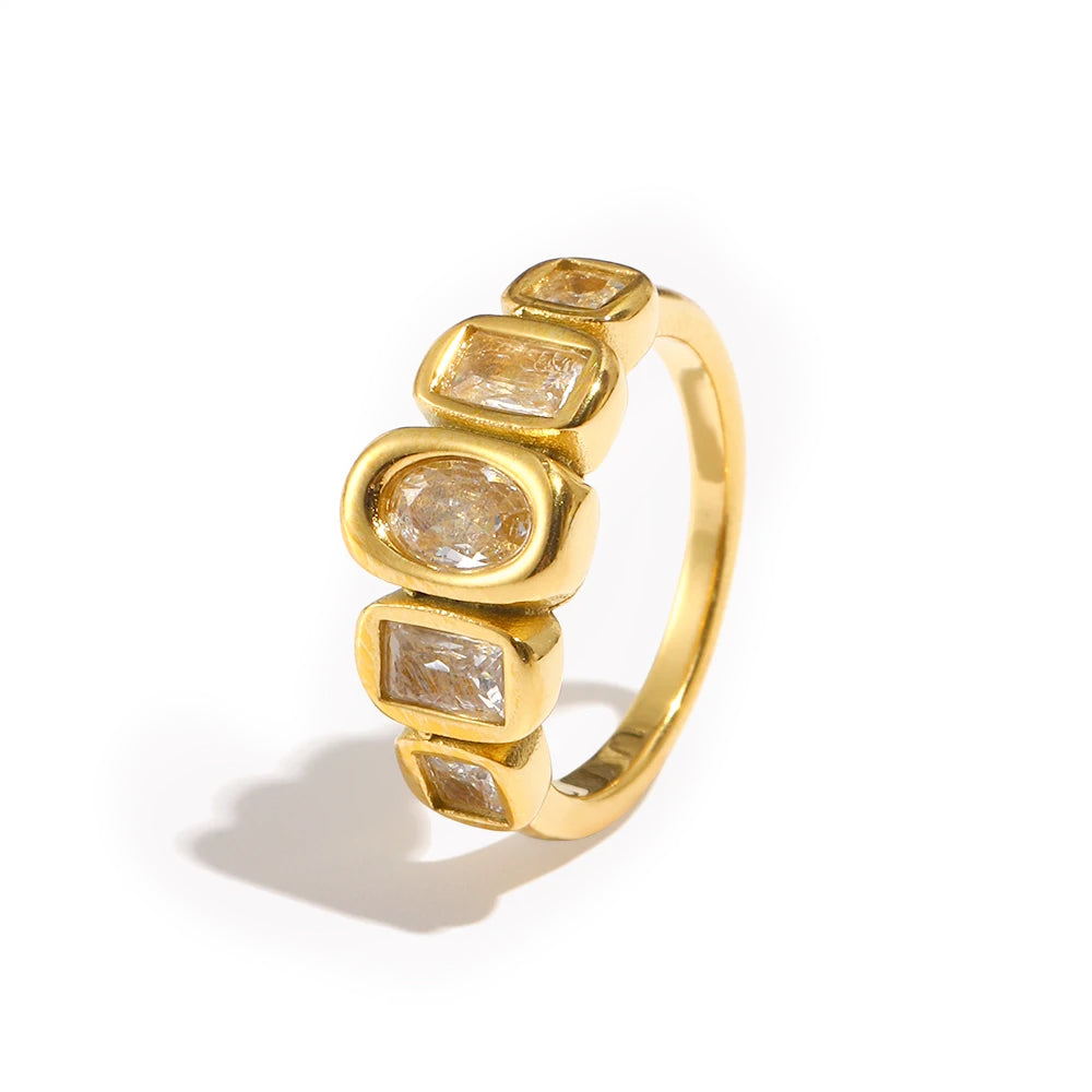 Gold oval and cushion multi-stone ring on white background, bezel-set oval center stone flanked by four cushion-cut accent stones in stepped formation, fine band with warm directional shadow
