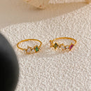 Two gold geometric multi-stone rings laid flat on textured white stone surface, tonal ring on left with deep green and clear baguette stones, multicolor ring on right with olive green, pink and clear stones, black stone and dark background

