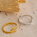 Gold and silver marquise stone snake rings laid flat on cream textured stone surface, back view showing tapered tail ends and open band gaps of both rings, smooth curved bands, yellow sandstone block in background, natural light
