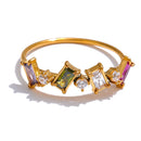 Gold geometric multi-stone ring on white background, close-up of multicolor colorway showing purple, olive green and clear baguette stones with round accent stones, prong settings on fine gold band, warm light
