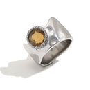 Wavy wide band crystal circle ring in silver, amber round stone with pavé surround on sculptural band, white background
