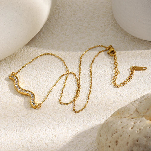 Gold pavé wave bar necklace laid flat on cream textured stone surface, full chain showing lobster clasp and extender, rhinestone wave pendant on left, white rounded stone objects in corners
