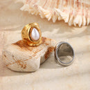 Gold hammered pearl chunky ring standing upright on pink stone at beach with silver version lying beside showing inner band, white shell in upper right corner, warm sandy background
