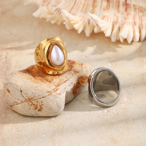 Gold hammered pearl chunky ring standing upright on pink stone at beach with silver version lying beside showing inner band, white shell in upper right corner, warm sandy background
