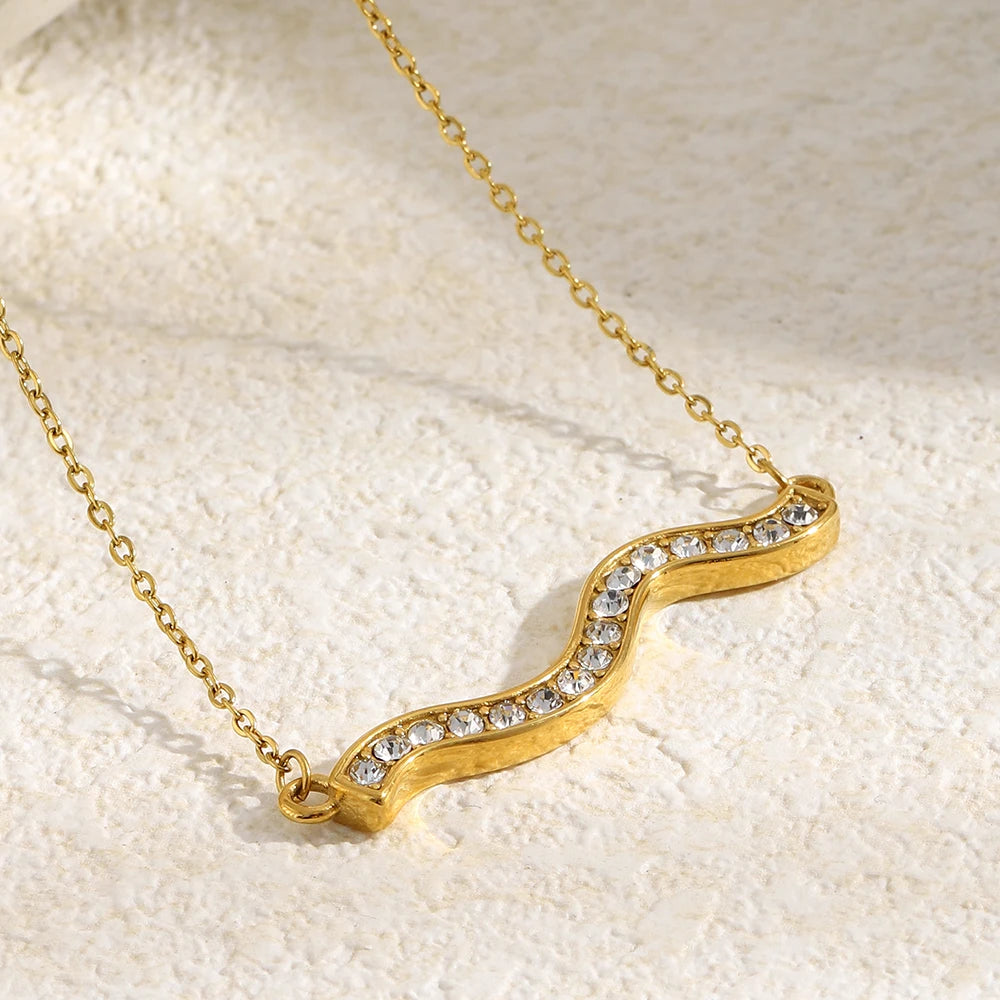 Close-up of gold pavé wave bar necklace pendant on cream textured fabric, S-curve wave silhouette with edge-to-edge rhinestone setting and gold border in sharp detail, white fabric in upper left corner
