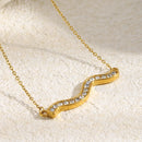 Close-up of gold pavé wave bar necklace pendant on cream textured fabric, S-curve wave silhouette with edge-to-edge rhinestone setting and gold border in sharp detail, white fabric in upper left corner
