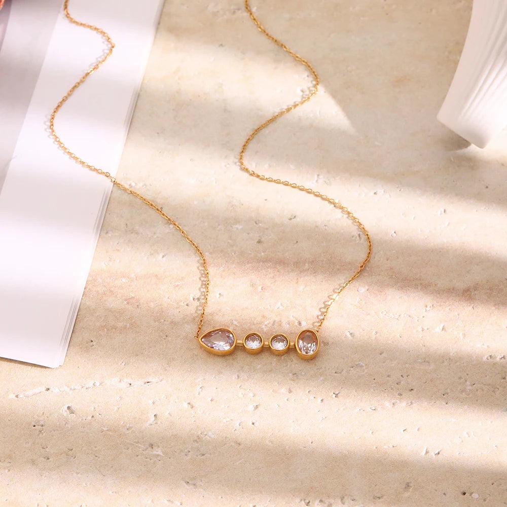 Gold graduated bezel stone bar necklace laid flat on cream stone surface, teardrop and round stones visible, white paper background, warm directional shadow
