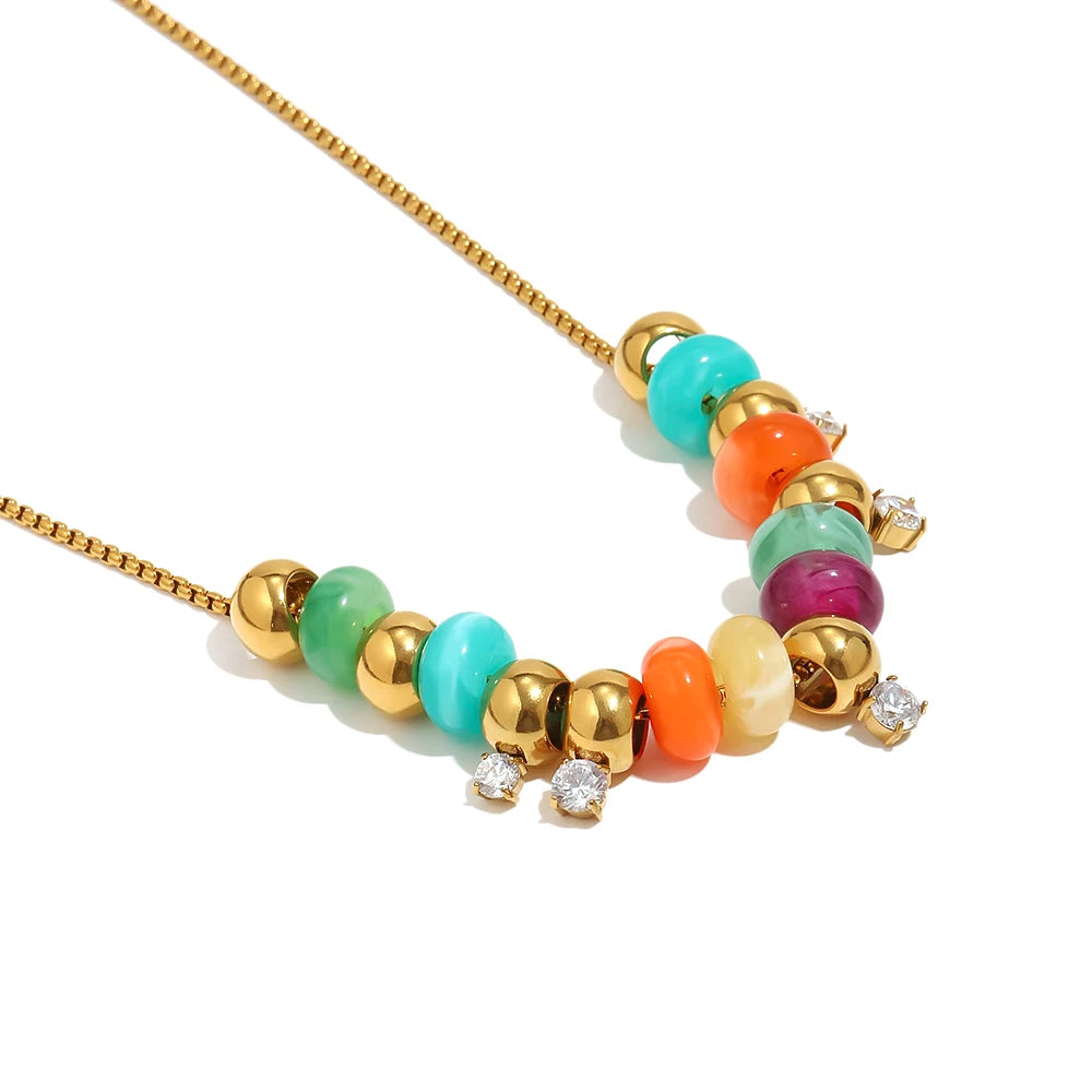 Close-up of gold colorful resin bead station necklace on white background, multicolor resin beads in turquoise coral amber and plum alternating with gold spheres, three clear stone stations visible, warm directional shadow
