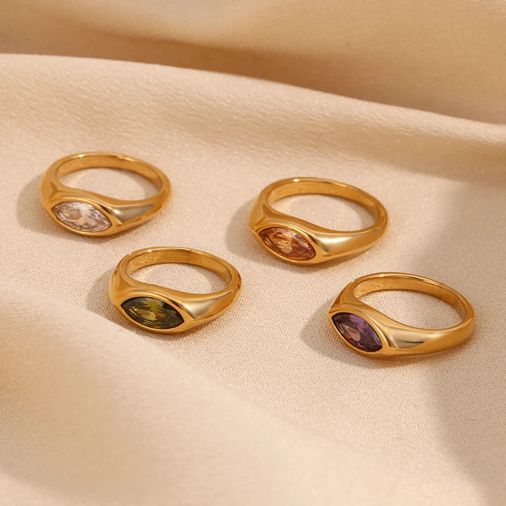 Four gold marquise stone dome signet rings scattered on cream velvet surface, white crystal, brown amber, green, and purple marquise stones in polished dome settings, soft warm ambient light
