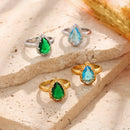 Four teardrop stone halo cocktail rings arranged on cream travertine stone surface, silver band versions with green and blue stones on upper level, gold band versions with green and blue stones on lower level, dried wheat stems in upper right corner
