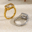 Gold emerald cut stone bezel ring face-up beside silver version showing back of setting on cream textured surface, gold bezel and step-cut stone in foreground, silver inner band and bezel base visible behind
