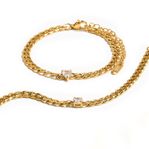 Two gold Cuban link bracelets on white background, upper bracelet with heart-cut stone and lower with round stone prong-set into curb chain links, lobster clasps and extender chains visible, warm shadow
