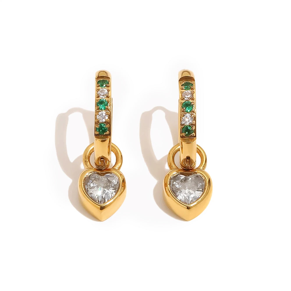 Gold huggie hoop earrings with alternating green and white pavé stones and bezel-set heart charm drop on white background, warm directional shadow
