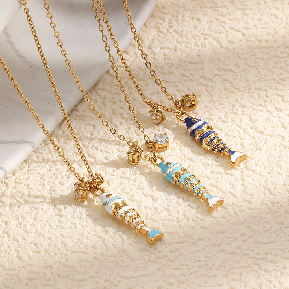 Three enamel fish pendant necklaces flat lay on cream textured surface, white blue and navy colorways
