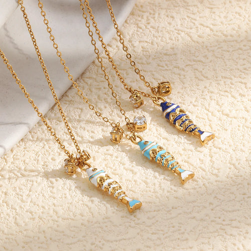 Three enamel fish pendant necklaces flat lay on cream textured surface, white blue and navy colorways

