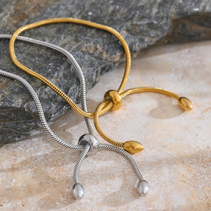 Gold and silver snake chain slider bracelets displayed together on marble surface, both showing barrel sliders and oval tips with fine snake chain texture, dark grey stone block in background, warm side light
