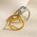 Gold and silver emerald cut stone bezel rings leaning against white cylindrical prop on cream textured surface, both showing stone face-up, bezel wall and band shoulder structure visible from above angle
