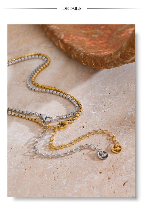 Gold and silver round stone tennis necklaces laid side by side on cream stone surface, both colorways showing continuous prong-set stones with lobster clasps, extender chains and engraved round tags, terracotta stone in background
