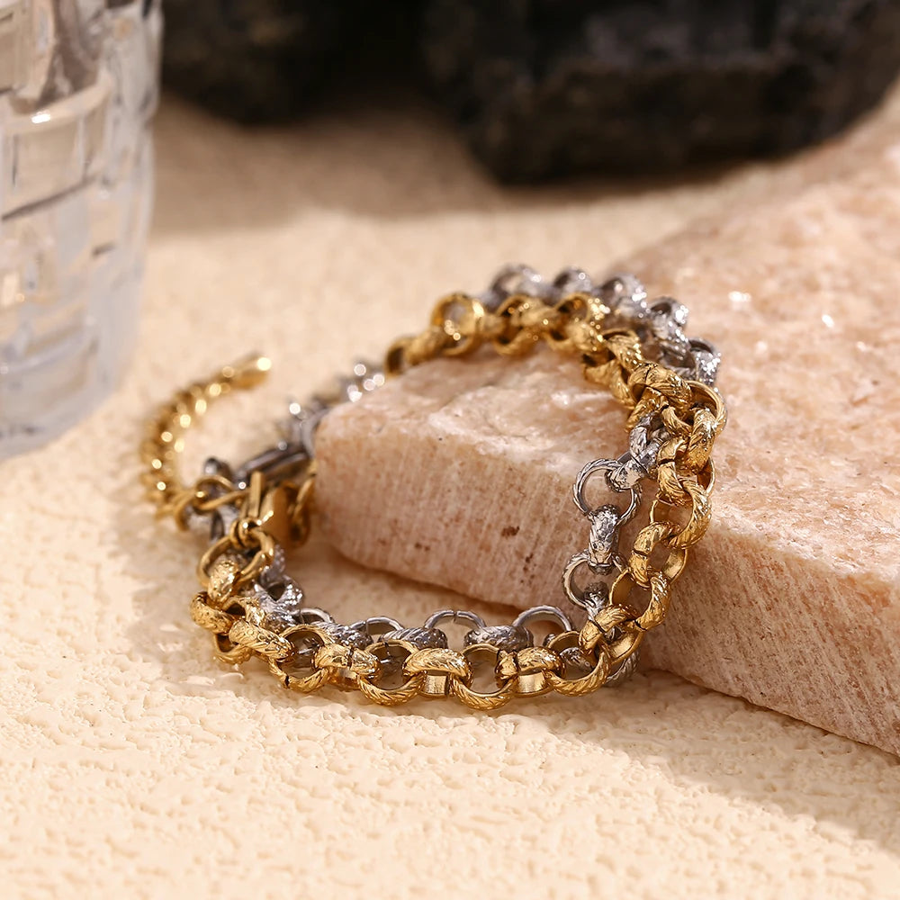 Gold and silver textured rolo chain bracelets draped over edge of cream stone block, side angle showing link thickness and hammered surface texture in warm light, dark stone and glass in background
