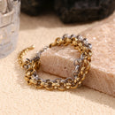 Gold and silver textured rolo chain bracelets draped over edge of cream stone block, side angle showing link thickness and hammered surface texture in warm light, dark stone and glass in background
