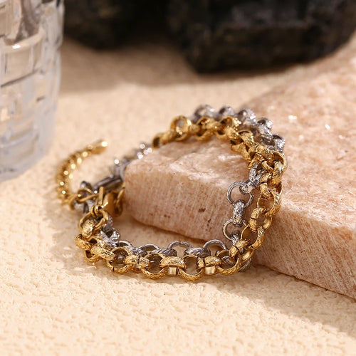 Gold and silver textured rolo chain bracelets draped over edge of cream stone block, side angle showing link thickness and hammered surface texture in warm light, dark stone and glass in background
