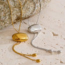 Gold and silver oval slider lariat necklaces laid side by side on cream textured stone surface, both showing polished dome oval sliders on fine snake chains with teardrop tips, rough grey stone in background, warm side light
