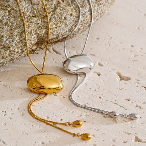 Gold and silver oval slider lariat necklaces laid side by side on cream textured stone surface, both showing polished dome oval sliders on fine snake chains with teardrop tips, rough grey stone in background, warm side light
