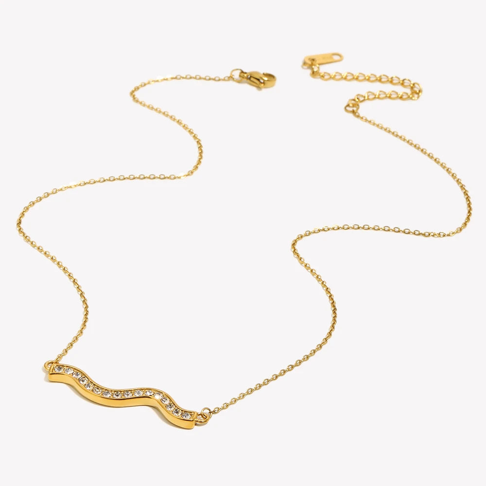 Gold pavé wave bar necklace on light grey-white background, overhead angled view showing full chain length with lobster clasp and extender at top, rhinestone wave pendant at bottom, fine O-chain links visible
