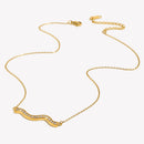 Gold pavé wave bar necklace on light grey-white background, overhead angled view showing full chain length with lobster clasp and extender at top, rhinestone wave pendant at bottom, fine O-chain links visible
